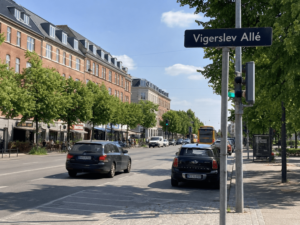 Vigerslev Allé i Valby med trafik, parkerede biler, butikker og etageejendomme